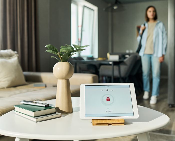 Tablet displaying smart home security interface standing on table in modern living room, Caucasian young adult woman entering background carrying bag, technology concept